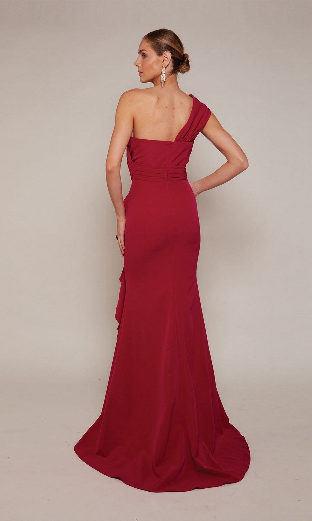 A woman wearing the Alyce 27646 long formal dress with a floor-length, one-shoulder sheath silhouette stands with her back to the camera against a plain background.